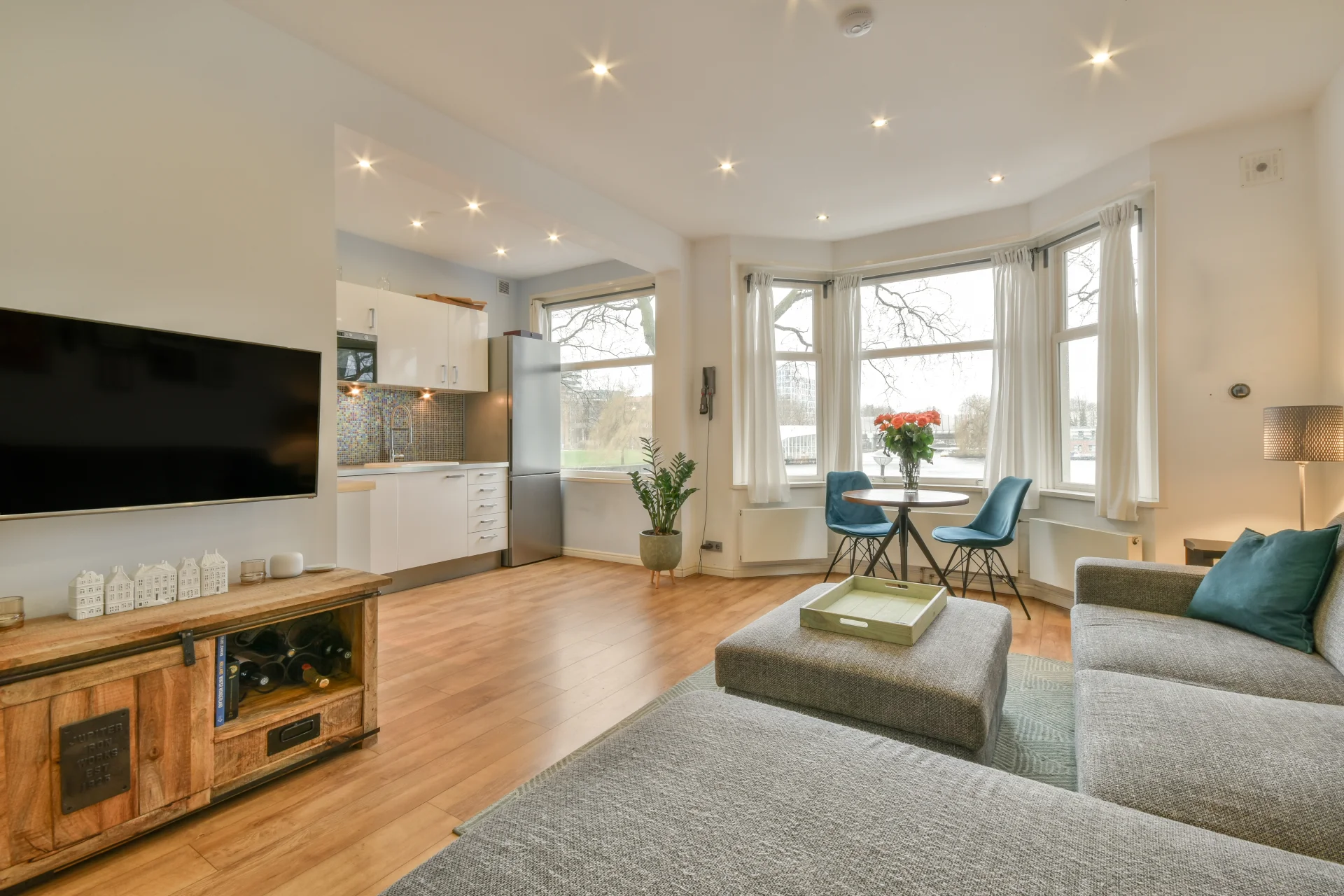Modern living room with a cozy gray sofa, a wooden coffee table, and a television, featuring bright natural light from large windows, a dining area with blue chairs, and a potted plant, emphasizing a supportive and structured sober living environment.