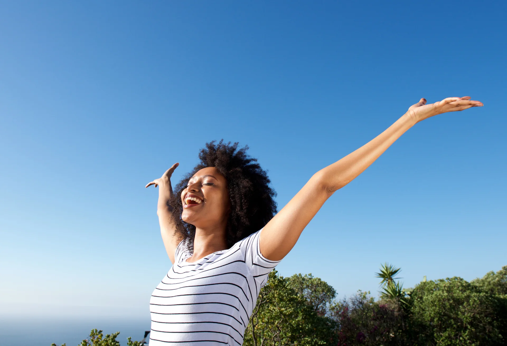 Woman with arms outstretched, smiling joyfully outdoors against a clear blue sky, symbolizing hope and freedom in recovery from addiction.